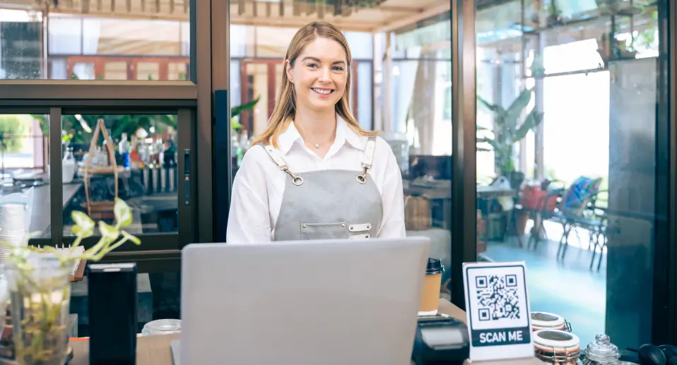 Restaurant staff member standing at the counter with a QR code sign beside a laptop in a bright modern cafe.